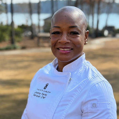 Chef Jestina Pambu smiling in her white chef's coat in front of Lake Oconee.