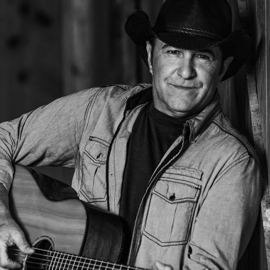 Tim Cadiere strums his guitar in this black and white headshot.